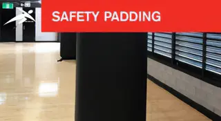 Black safety padding installed on a pole inside a sports hall with timber flooring. A red banner at the top reads “SAFETY PADDING” with HiTech’s logo.
