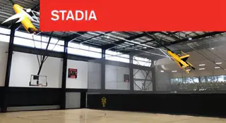 Modern indoor stadium with a basketball court, scoreboard, and large spectator seating area. A red banner at the top reads “STADIA” with HiTech’s logo.